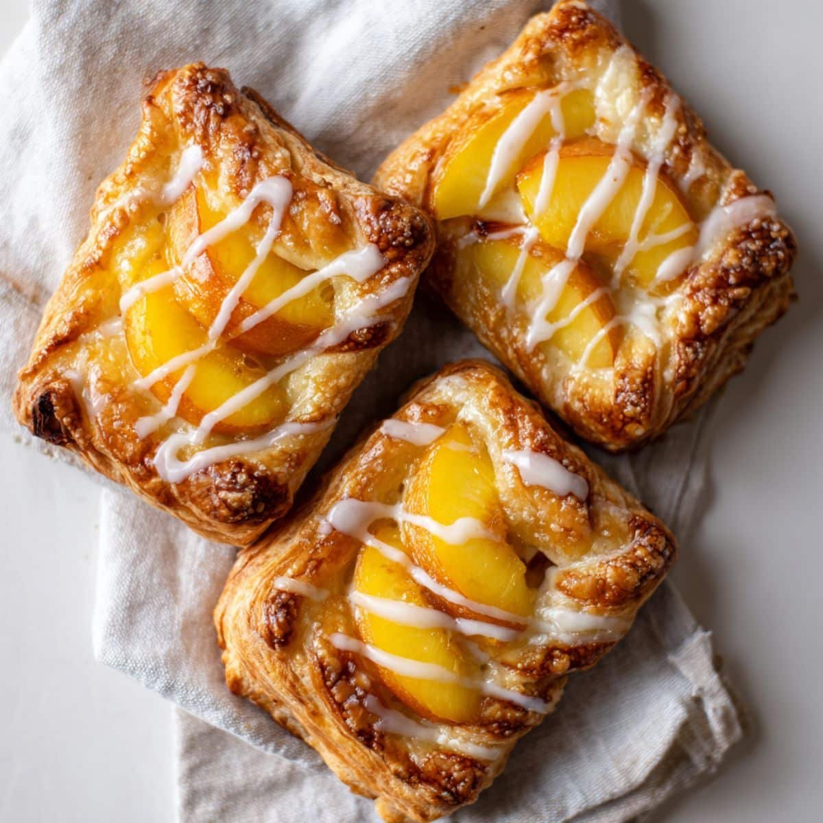Homemade Peach Danish Pastry on a white kitchen table, golden brown with peach slices and glaze, photographed from above.
