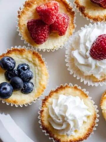 Homemade keto mini cheesecakes on a white kitchen table, shot from above, with simple toppings like berries and whipped cream.