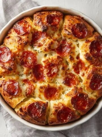 Pull-apart pepperoni bread recipe in a round baking dish, golden brown with melted cheese and pepperoni on a white kitchen table, top-down shot