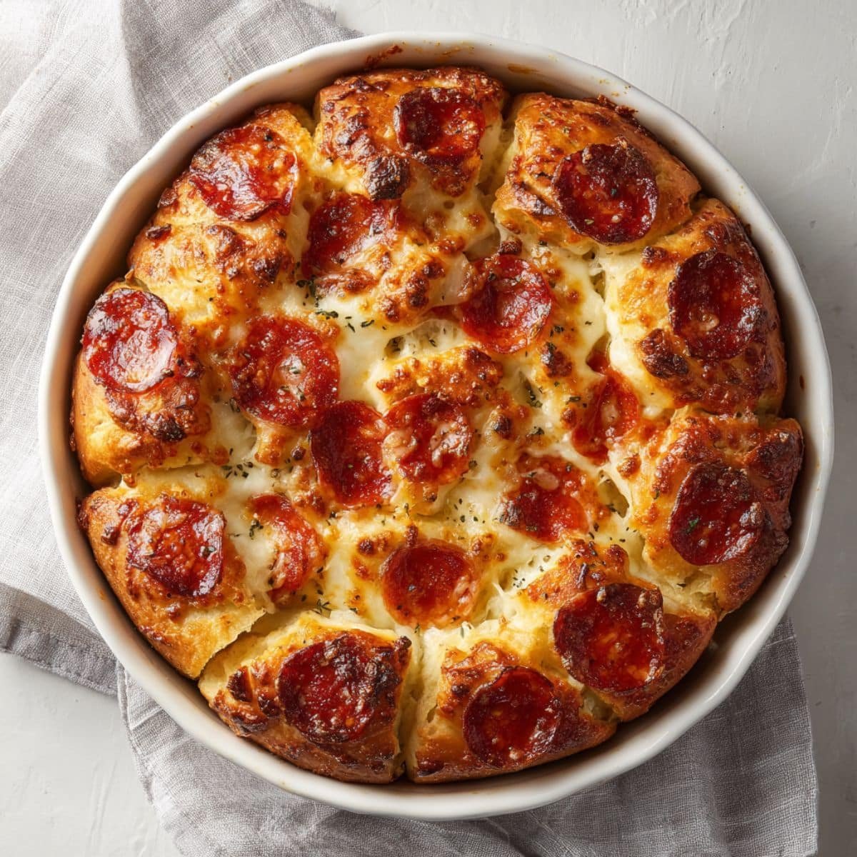 Pull-apart pepperoni bread recipe in a round baking dish, golden brown with melted cheese and pepperoni on a white kitchen table, top-down shot