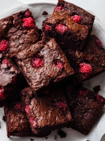 Top-down photo of fudgy raspberry brownies with fresh raspberries on parchment paper on a white kitchen table.