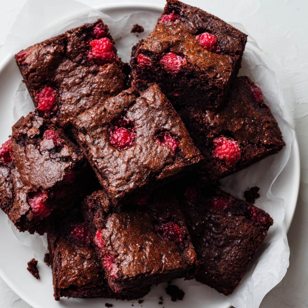 Top-down photo of fudgy raspberry brownies with fresh raspberries on parchment paper on a white kitchen table.