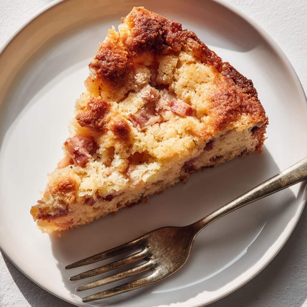 A rustic slice of homemade rhubarb cake on a white plate, photographed from above on a white kitchen table.