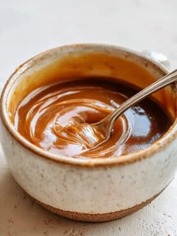 Homemade vegan caramel sauce in a glass jar with a spoon, photographed from above on a white kitchen table.