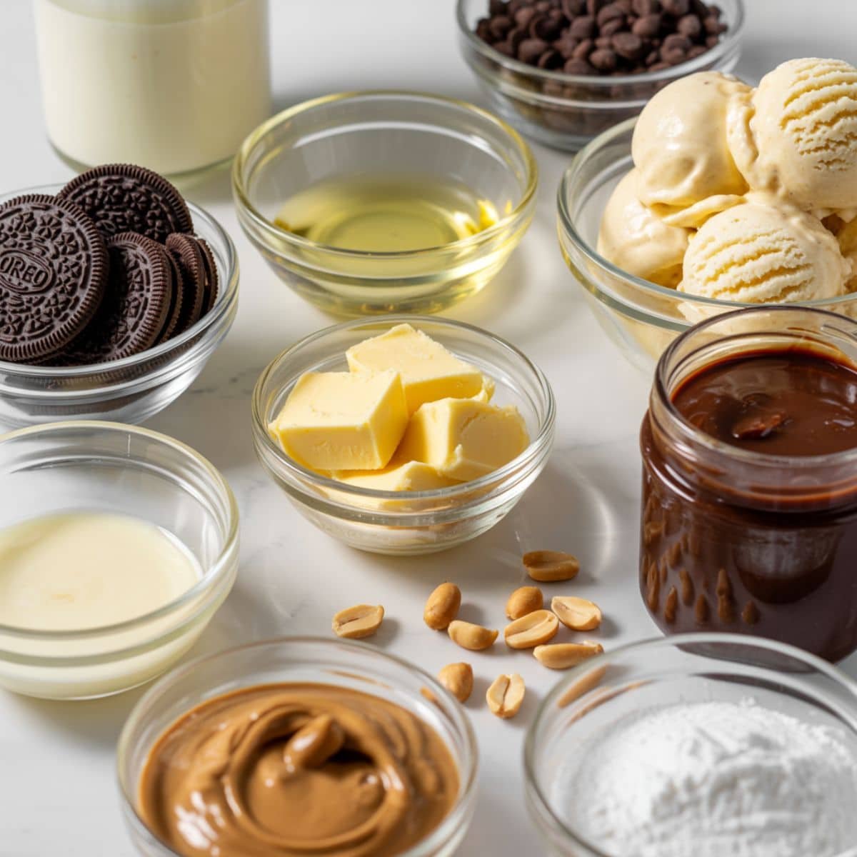 All ingredients for a Buster Bar Ice Cream Cake — Oreos, butter, vanilla ice cream, fudge sauce, peanuts, chocolate chips, coconut oil, peanut butter, cream, and sugar on a white kitchen counter.