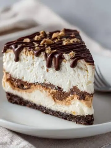 A homemade Buster Bar Ice Cream Cake slice showing Oreo crust, creamy vanilla ice cream, fudge layer, and roasted peanuts on a white kitchen table.