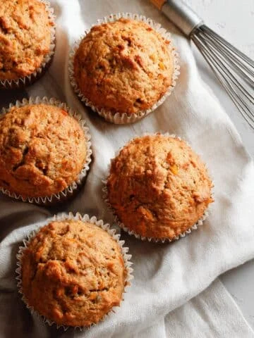Freshly baked homemade carrot muffins cooling on a white kitchen table with a whisk and linen towel nearby, shot from above in natural light.