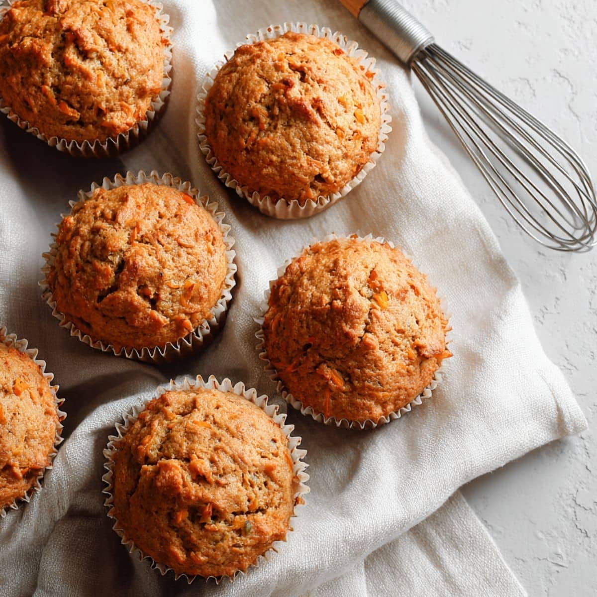 Freshly baked homemade carrot muffins cooling on a white kitchen table with a whisk and linen towel nearby, shot from above in natural light.