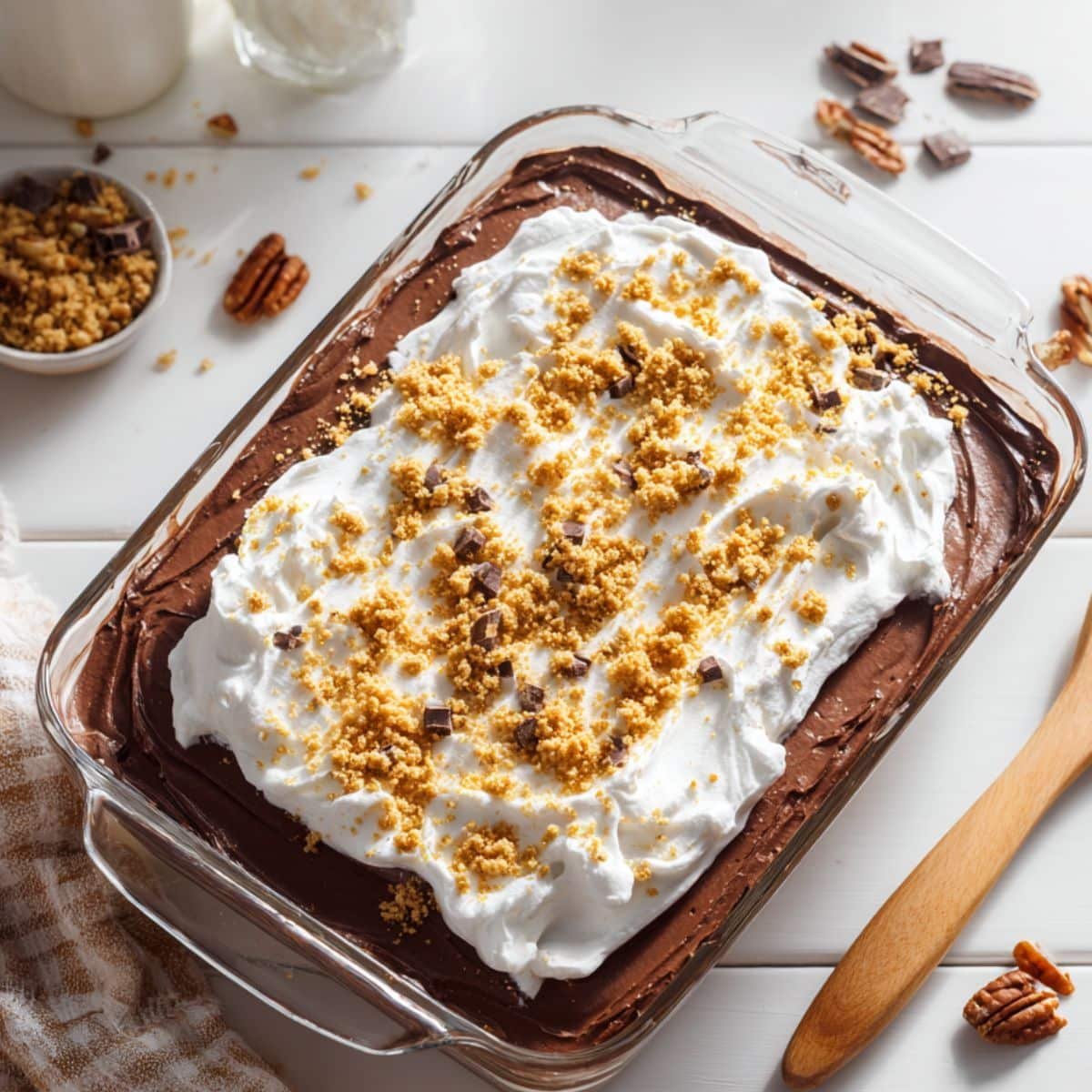 A homemade layered chocolate delight recipe in a glass dish on a white kitchen table, topped with whipped cream, chocolate shavings, and pecans.