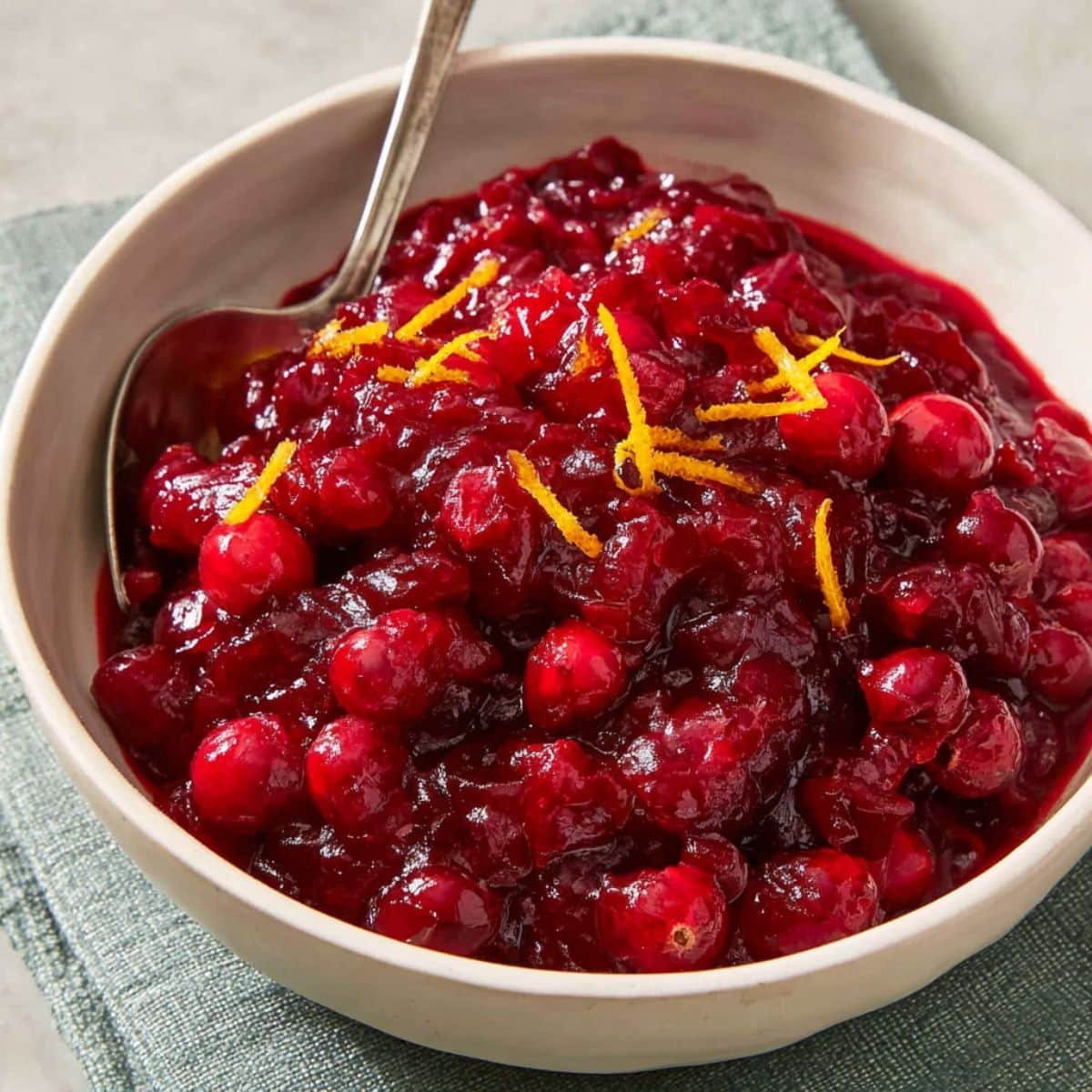 Homemade cranberry sauce with fresh cranberries and orange zest served in a white bowl on a white kitchen table, photographed from above.