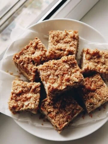 Top-down photo of homemade apple oatmeal breakfast bars on parchment paper over a white kitchen table, with a few crumbs and soft natural light.