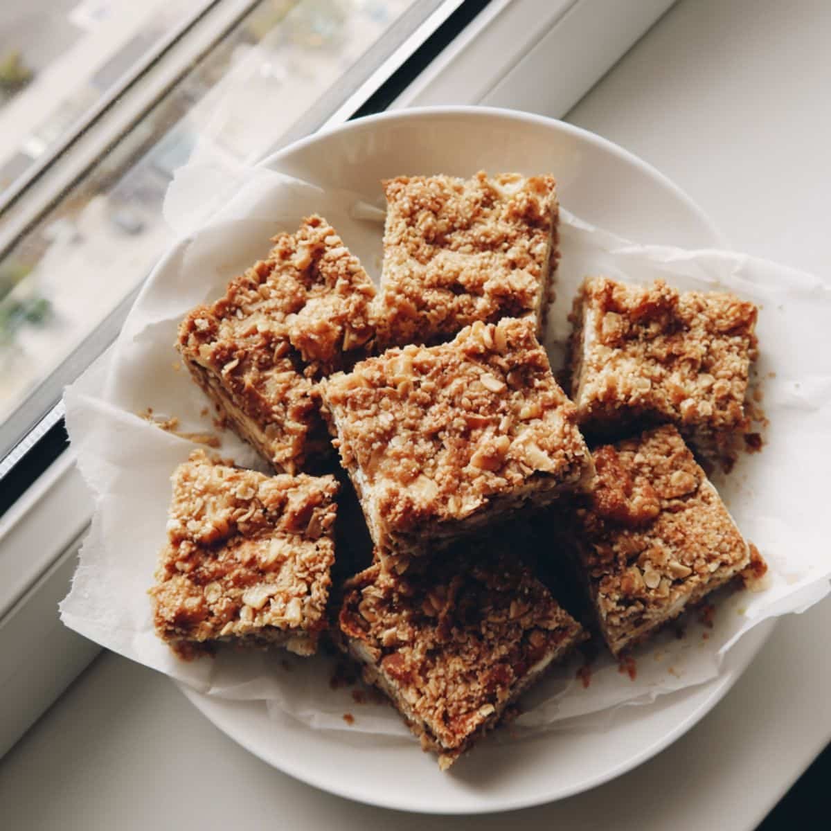 Top-down photo of homemade apple oatmeal breakfast bars on parchment paper over a white kitchen table, with a few crumbs and soft natural light.