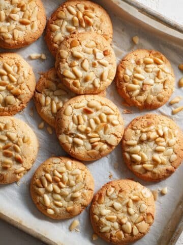 Freshly baked Italian pignoli cookies topped with golden pine nuts, cooling on parchment paper over a white kitchen counter, captured from above in natural light.