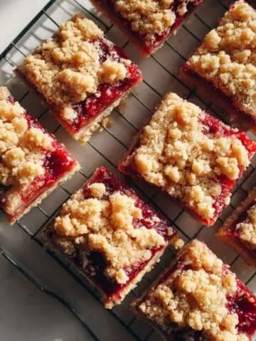 Freshly baked raspberry bars with golden crumb topping and raspberry filling cooling on a white kitchen counter.