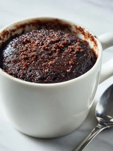 Fudgy brownie mug cake in a white mug on a kitchen table with a spoon beside it, shot from above.