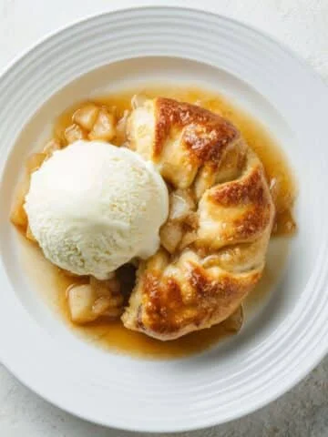 Freshly baked homemade apple dumplings in a white baking dish with buttery cinnamon sauce on a white kitchen counter