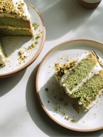 Homemade green cake slice on a white kitchen table, topped with creamy frosting and pistachio crumbs, photographed from above in natural light.