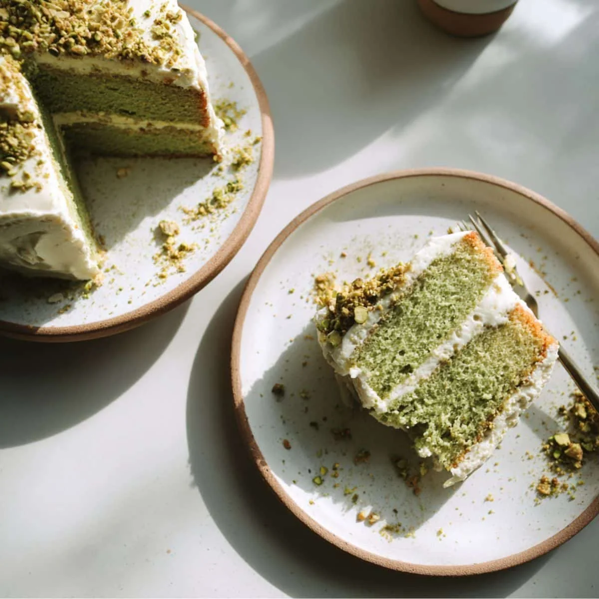 Homemade green cake slice on a white kitchen table, topped with creamy frosting and pistachio crumbs, photographed from above in natural light.