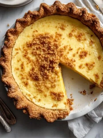 Homemade egg custard pie slice on a white plate, showing creamy filling and flaky crust on a white kitchen table.
