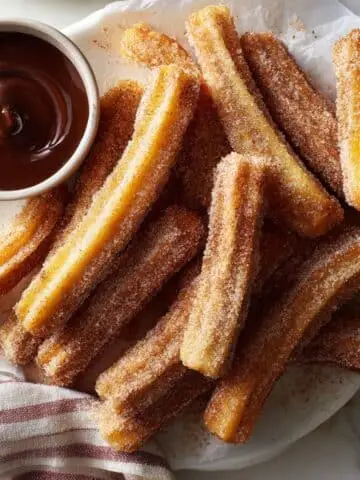 Homemade gluten-free churros dusted with cinnamon sugar on a white kitchen table, served with chocolate sauce.