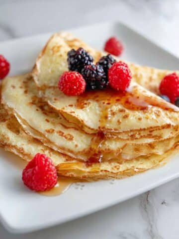 Homemade keto crepes recipe stacked on a white kitchen counter with berries and syrup.