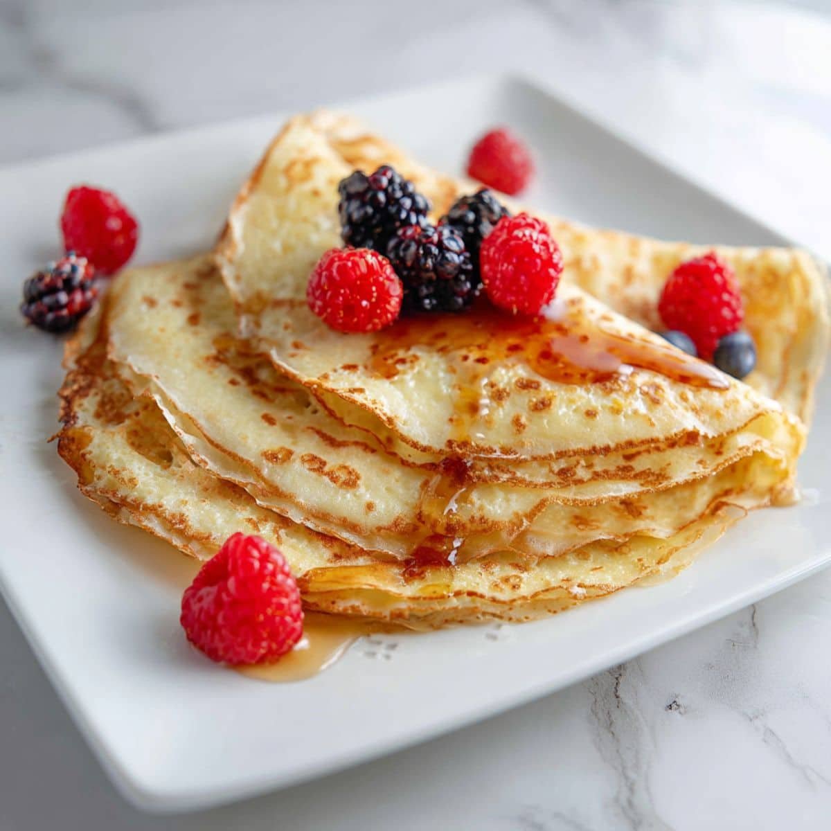 Homemade keto crepes recipe stacked on a white kitchen counter with berries and syrup.