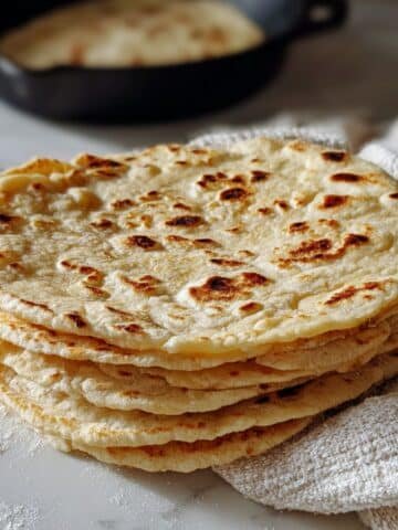 Stack of freshly made sourdough tortilla recipe on a white kitchen counter with flour dust and a cast iron skillet in the background.