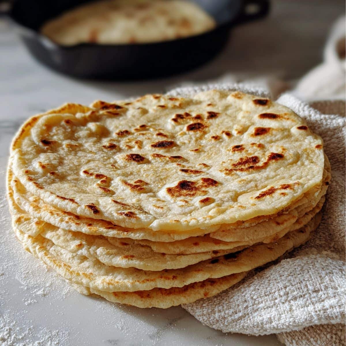 Stack of freshly made sourdough tortilla recipe on a white kitchen counter with flour dust and a cast iron skillet in the background.