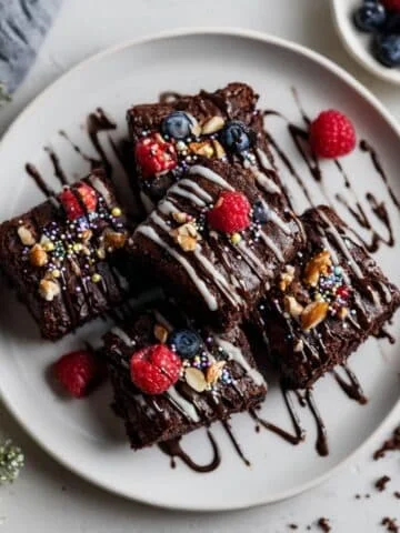 Homemade chocolate brownies decorations with ganache, nuts, and sprinkles on a white kitchen table, shot from above in natural daylight.