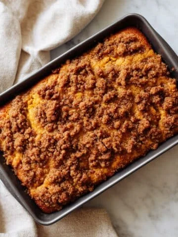 Homemade pumpkin cinnamon coffee cake with streusel topping on a white kitchen table, sliced imperfectly to show moist pumpkin texture.