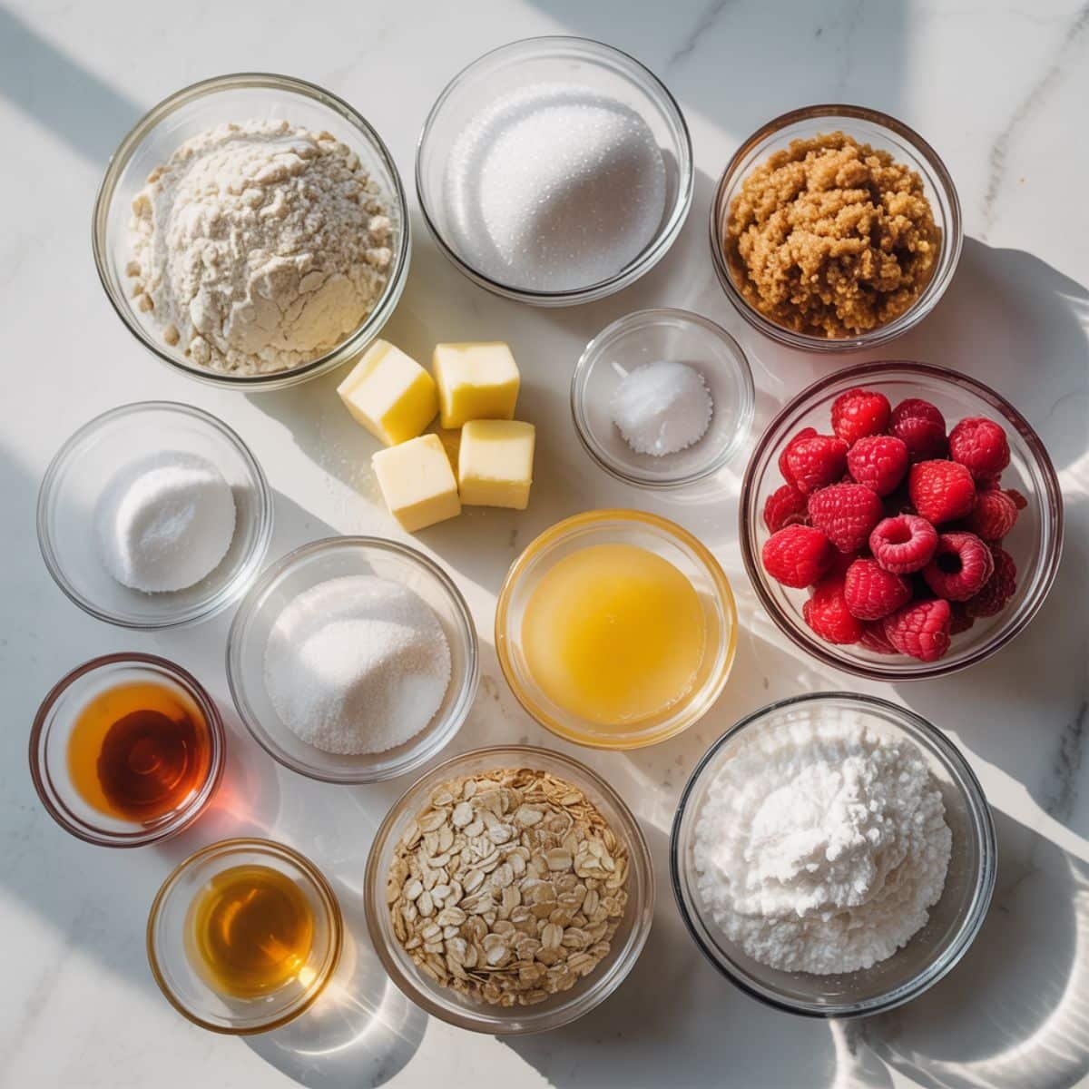 Ingredients for raspberry bars — flour, sugar, butter, raspberries, lemon juice, oats, and cornstarch arranged on a white kitchen table.
