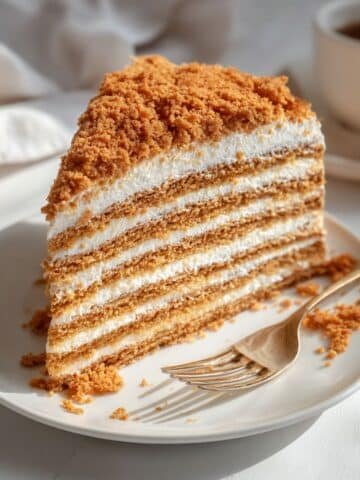 Top-down photo of a homemade Russian honey cake slice showing creamy layers and honey crumbs on a white kitchen table.