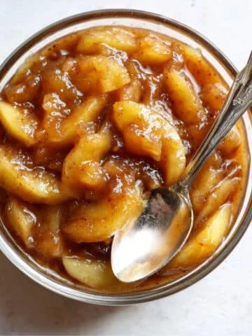 Homemade apple pie filling in a glass bowl with cinnamon sauce on a white kitchen counter, photographed from above.