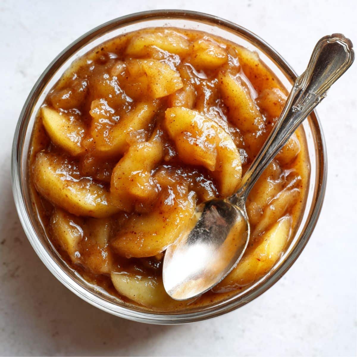 Homemade apple pie filling in a glass bowl with cinnamon sauce on a white kitchen counter, photographed from above.