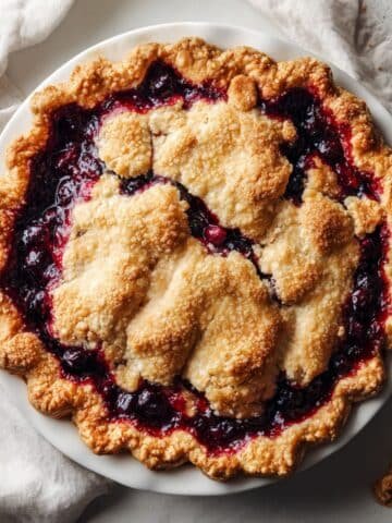 Freshly baked homemade Saskatoon pie with golden crust on a white kitchen table, crumbs and juice bubbling over, captured in natural light.