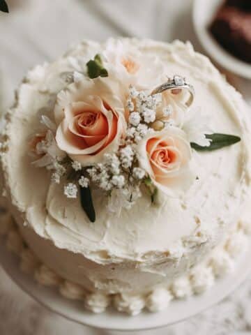 Homemade engagement cake with white buttercream and fresh flowers, shot from above on a white kitchen table in natural light.