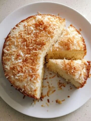 Homemade vegan coconut cake topped with shredded coconut, sliced on a white plate, photographed from above on a kitchen table.