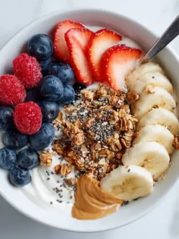 Homemade granola bowl with yogurt, fresh berries, banana slices, granola, and honey on a white kitchen table