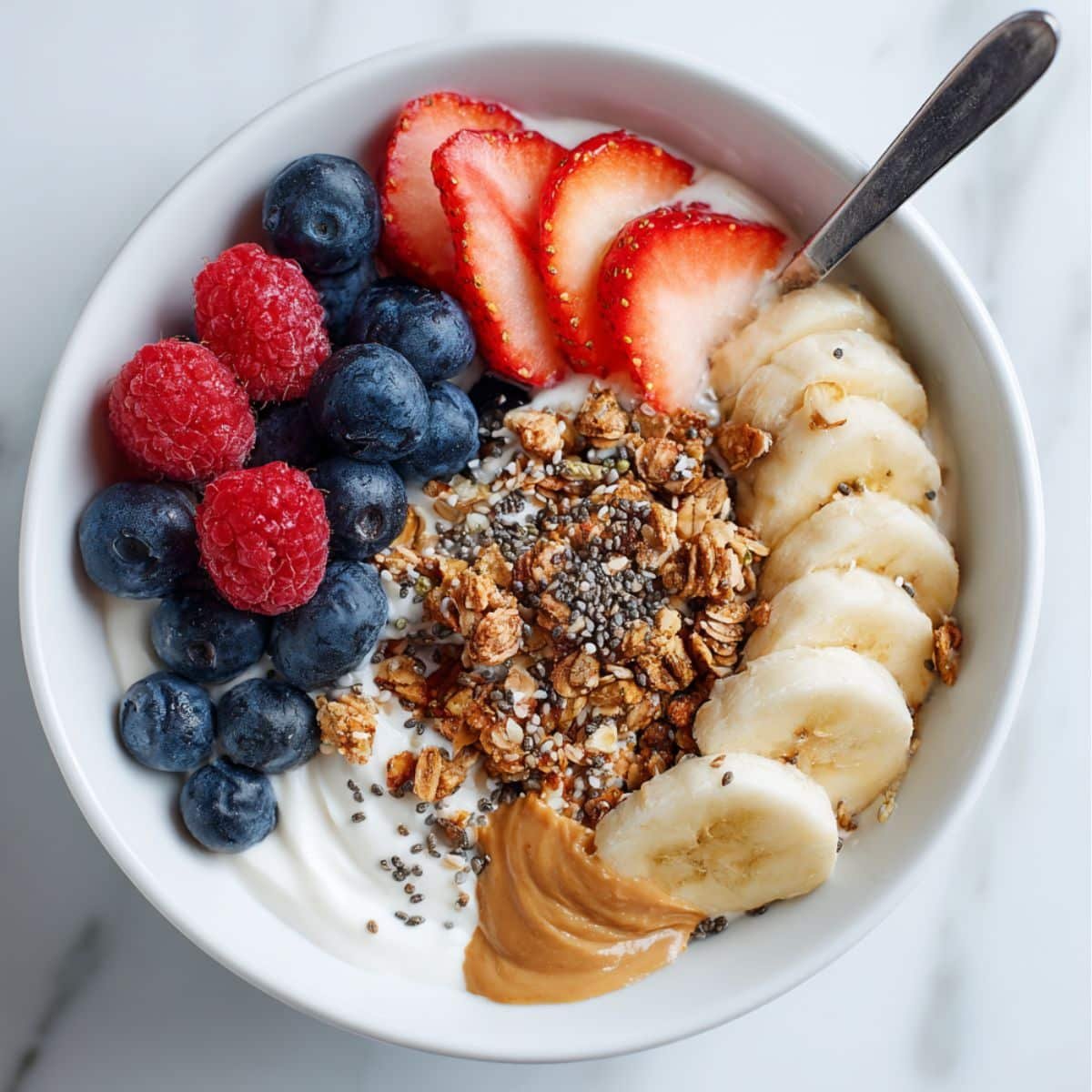 Homemade granola bowl with yogurt, fresh berries, banana slices, granola, and honey on a white kitchen table