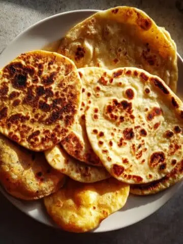 Assorted homemade Indian breads recipes— naan, roti, paratha, and poori — on a white kitchen table, photographed from above with natural light.