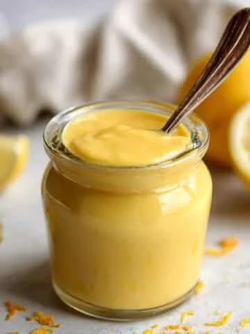 Homemade vegan lemon curd in a glass jar with a spoon on a white kitchen counter.