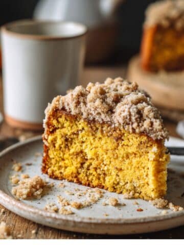 Vegan pumpkin coffee cake slice on a white plate with crumb topping and coffee cup in natural light.