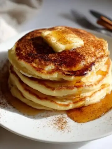 Fluffy cinnamon pancakes stacked on a white plate, topped with butter and syrup, photographed from above with a cozy homemade vibe.