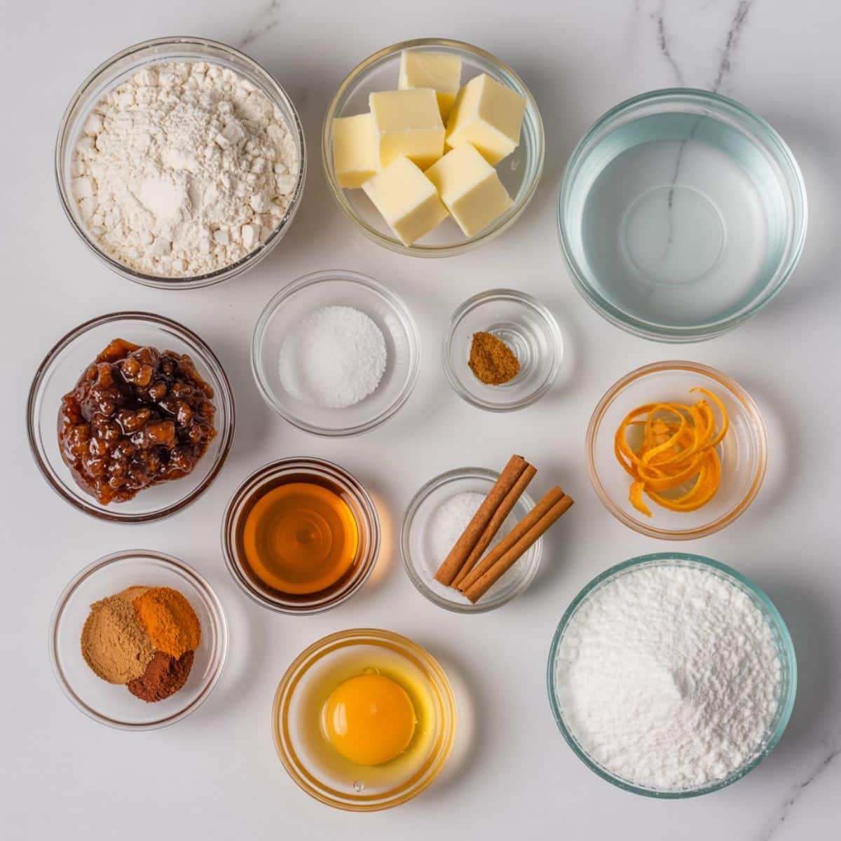 Flat-lay of gluten free mince pie ingredients in small bowls on a white counter with butter cubes, flour, spices, and orange zest.