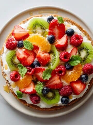 Homemade healthy fruit pizza with fresh colorful fruits on a white plate, photographed from above on a kitchen counter.