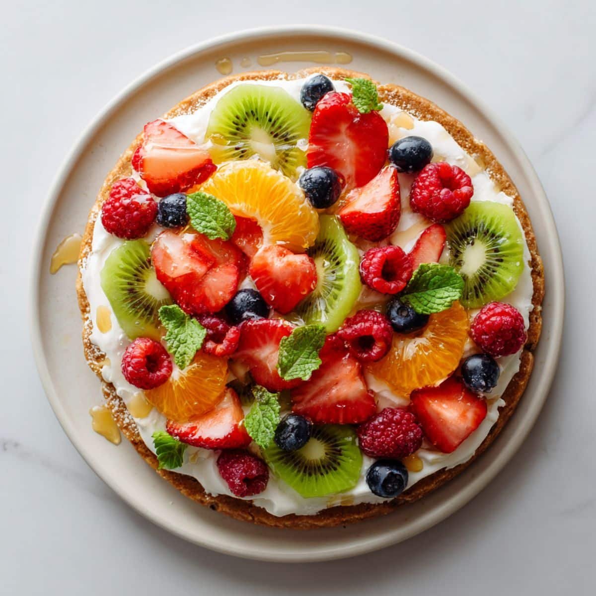 Homemade healthy fruit pizza with fresh colorful fruits on a white plate, photographed from above on a kitchen counter.