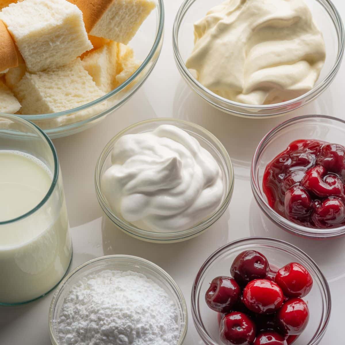 All ingredients for a homemade heaven on earth cake — angel food cake cubes, pudding mix, milk, Cool Whip, cherries, and powdered sugar on a white countertop.